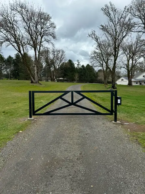 Black double-swing automatic driveway gate with operator across gravel farm drive