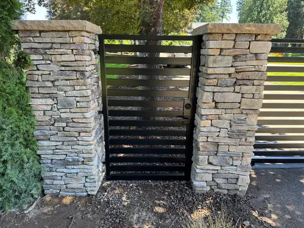 Black horizontal-slat pedestrian gate with lock between stacked-stone pillars, matching the main driveway gate